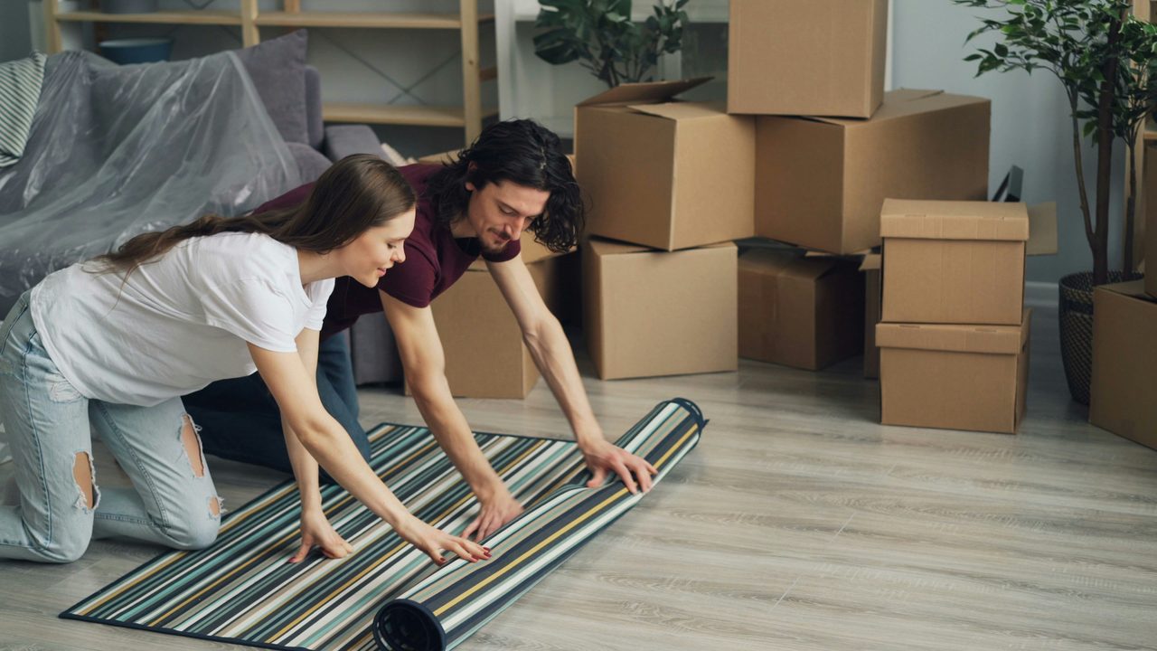 A couple unrolling a rug in a new apartment amid stacked moving boxes