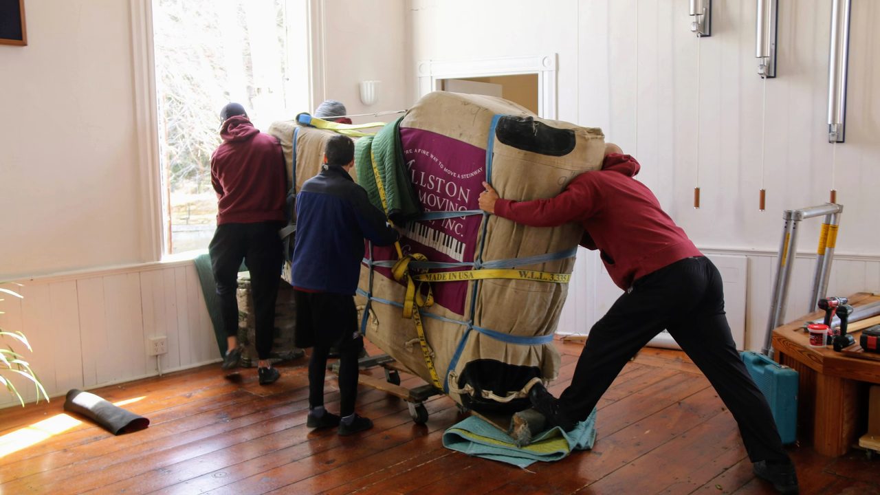 A crew easing a padded, strapped piano across a room on a dolly